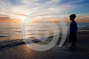 Silhouette of boy in front of a sunset, looking at the ocean