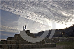 The silhouette of a boy and a girl on top of the bale of hay group in a pyramid wall pattern in agriculture farm at sunset