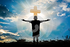 Silhouette of boy praying to a cross with heavenly cloudscape sunset concept for religion, worship, love and spirituality