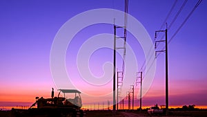 Silhouette of bulldozer tractor with row of electric poles in countryside area against colorful twilight sky background