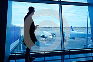 Silhouette of businessman in airport lounge with mobile phone standing by panoramic windows overlooking runways and airplanes