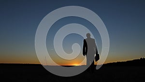 Silhouette of businessman with briefcase walking across field at sunset, against a blue sky