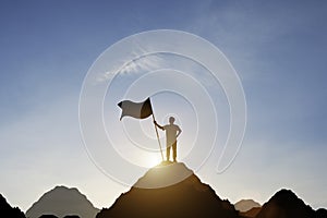 Silhouette of businessman holding flag on the top of mountain with over blue sky and sunlight. It is symbol of leadership