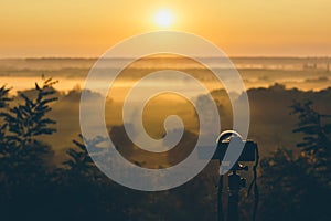 Silhouette of a camera on a tripod on a hill capturing sunrise and morning fog. Countryside landscape with camera on tripod,