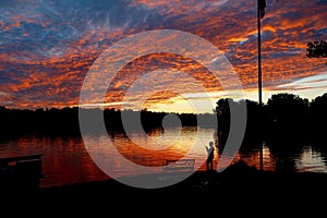 Silhouette of child fishing in a lake during a fiery, summer sunset.