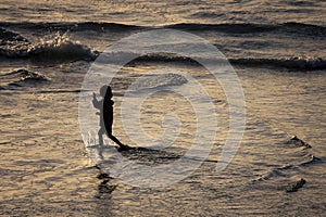 A silhouette of a child playing on a beach at sunset with reflections on the wet sand and waves behind