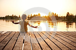 Silhouette of a child sitting on the wooden pier and enjoying heat summer evening at the lake at sunset