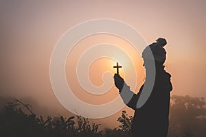 Silhouette of christian young woman praying with a cross at sun