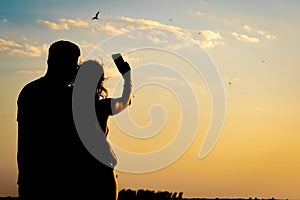 Silhouette of a couple embracing, taking a photo with their cell phone, of birds flying over the sea during a sunset