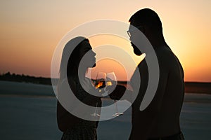 Silhouette of a couple with glasses on sunset background, man and woman clanging wine glasses with champagne at sunset dramatic sk