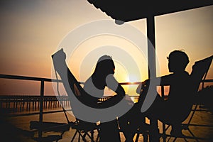 Silhouette of couple lover sitting and relax on romantic sunset at the sea of honeymoon vacation.