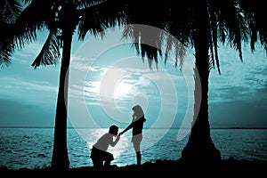 Silhouette couple, woman stand and man sit on the floor, hold and kissing her hand for propose her to wedding in front of the sea