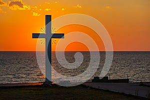 Silhouette of cross at sunrise or sunset with light rays and sea panorama