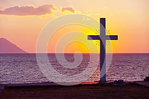 Silhouette of cross at sunrise or sunset with light rays and sea panorama