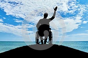 Silhouette of a disabled man in a wheelchair friendly waving his hand atop a hill near the sea