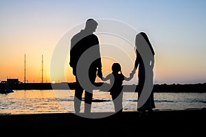 Silhouette of a family at the beach holding hands during sunset. Father and mother look at their daughter. Romantic and passionate