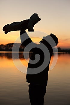 Silhouette of smiling father holding little baby in his handson sunset with lake on background. Man with child. Family concept. Fa