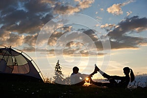 Silhouette of female and boy sitting on grass in yoga pose at daybreak near tent