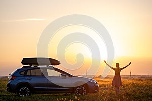 Silhouette of female driver standing near her car on grassy field enjoying view of bright sunset. Young woman relaxing