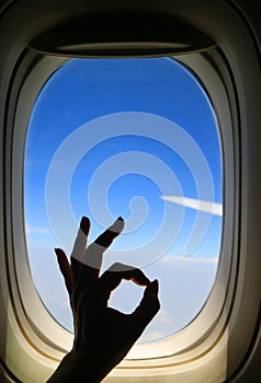 Silhouette of female`s hand posing OK sign against airplane window with vibrant blue sky and airplane wing