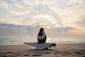 Silhouette Of Female Surfer At The Beach Sitting On Sand With Surfboard At Sunset