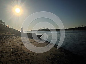 Silhouette of fisherman casting his fishing rod. Landscape of river and fishermen boats at sandy beach in summer morning