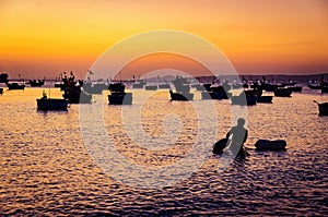 Silhouette of a fisherman cleaning his nets in the sea with fishing boats in the background in Mui Ne fishing village during