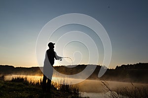 Silhouette of the fisherman at dawn catching fish in the river. Cold summer morning and mist over the river.