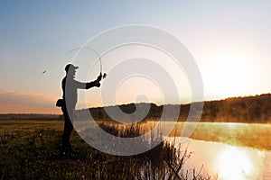 Silhouette of the fisherman at dawn catching fish in the river. Cold summer morning and mist over the river.