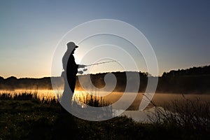 Silhouette of the fisherman at dawn catching fish in the river. Cold summer morning and mist over the river. photo