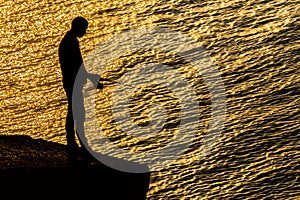 The silhouette of a fisherman with a fishing rod in front of glittering sea water.