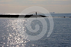 Silhouette of a fisherman on the rocky cliff, at sunset, with a ship on the horizon, Fiumicino, Rome, Lazio, Italy