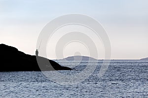 Silhouette of a Fisherman standing on rocks with fishing rod near sea on Sunny day