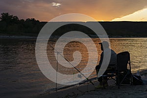 Silhouette of fisherman in sunset sitting on bank of river with fishing rod in summer evenong and looking over river,