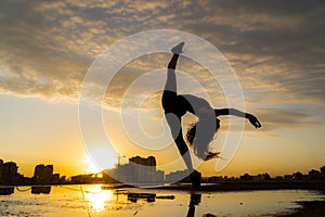 Silhouette Flexible female dancer making trick with reflection in the water during dramatic sunset. Concept of happiness