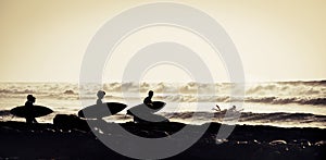 Silhouette of four surfers entering at the water with surfboards and wetsuits - surfing at the sunset and with waves at the