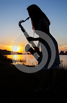 Silhouette of a girl in a dress at dawn near the river in the village playing saxophone on a background of blue sky