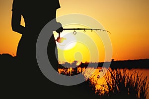 Silhouette of a girl with a fishing rod on the river bank at dawn