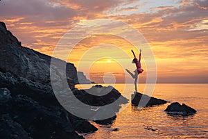 The silhouette girl on a rocky beach, against the dawn, in the pose of a jumping ballerina on stage, with outstretched wings in