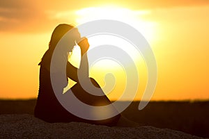 Silhouette of girl sitting on the sand and praying to God at sunset, the figure of young woman on the beach, concept religion