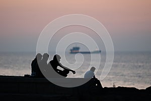 The silhouette of a group of unidentified people sitting on the embankment against the sea at sunset and a passing ship in the