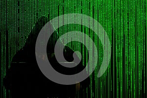 Silhouette of a hacker in a hood with a laptop on a background of a matrix of binary computer code. Concept: hacker and computer