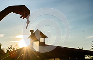 Silhouette of a hand holding a black miniature house model and house keys against background of evening sky and sunset there are