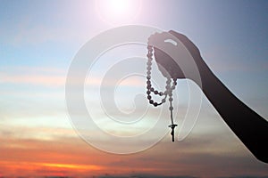 Silhouette of a hand holding Rosary on colorful sunset sky light background. Person with Rosary in hands with Jesus Christ