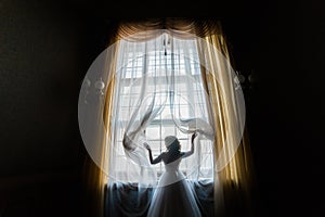 Silhouette of happy bride in white dress opening curtains