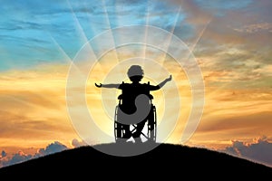 Silhouette of a happy disabled child girl sitting in a wheelchair atop a hill at sunset photo