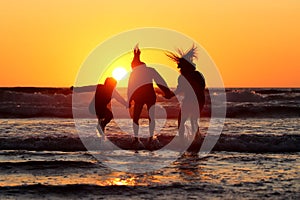 Silhouette of happy friends holding hands and jumping in the water on a coastline at sunset