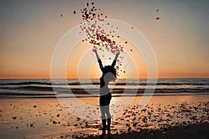Silhouette of a happy woman with open arms free on a beach, throwing red rose petals into the air..