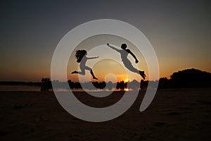 Silhouette of a happy young couple having fun outdoors, man and woman jumping on the beach by the river, lake on sunset