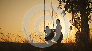 Silhouette of happy young mother and little daughter on a swing at sun light. Pretty girl sitting on a wooden swing and